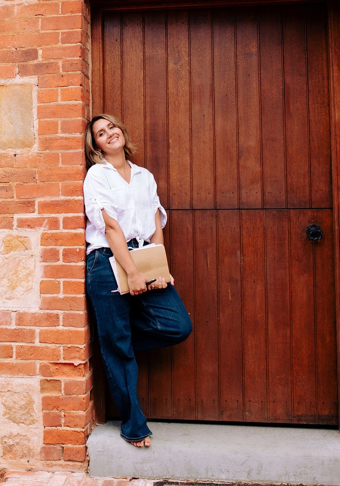 Renée Melizza, Adelaide celebrant, Poet and Author, leaning against a brick doorway with her journals in hand, photo by Kirsty Caruso Photography