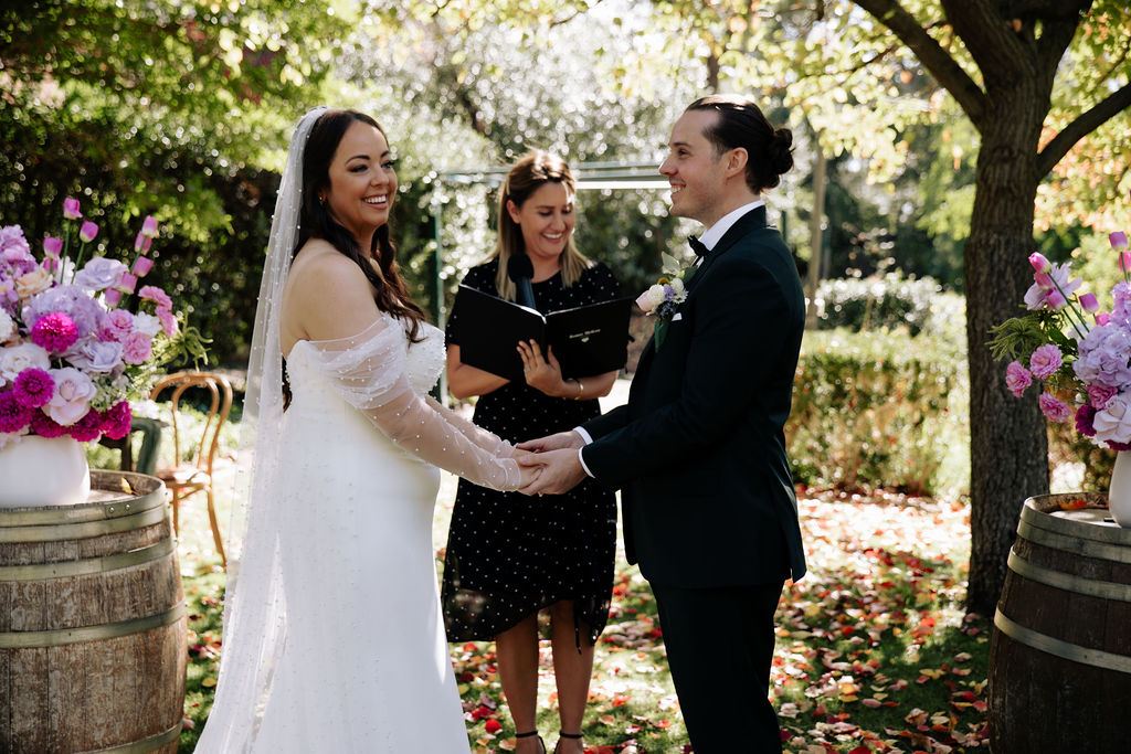 Renée Melizza officiating as a bride and groom hold hands and laugh during a garden wedding ceremony, photo by Kirsty Caruso Photography