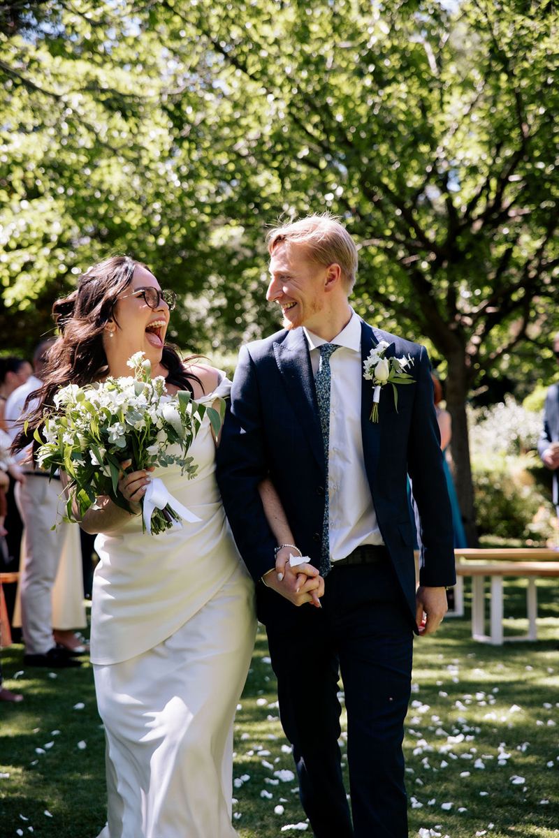Luiza and Matt laughing together as they walk down a petal-covered garden aisle, photo by Kirsty Caruso Photography