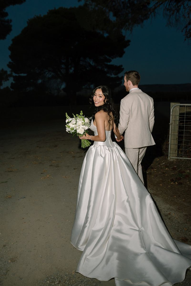 Jeongah glancing back with her bouquet as she and Anthony walk hand in hand at dusk