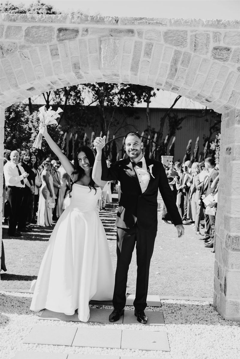 Joanna and Michael celebrating with arms raised beneath a stone archway as guests applaud