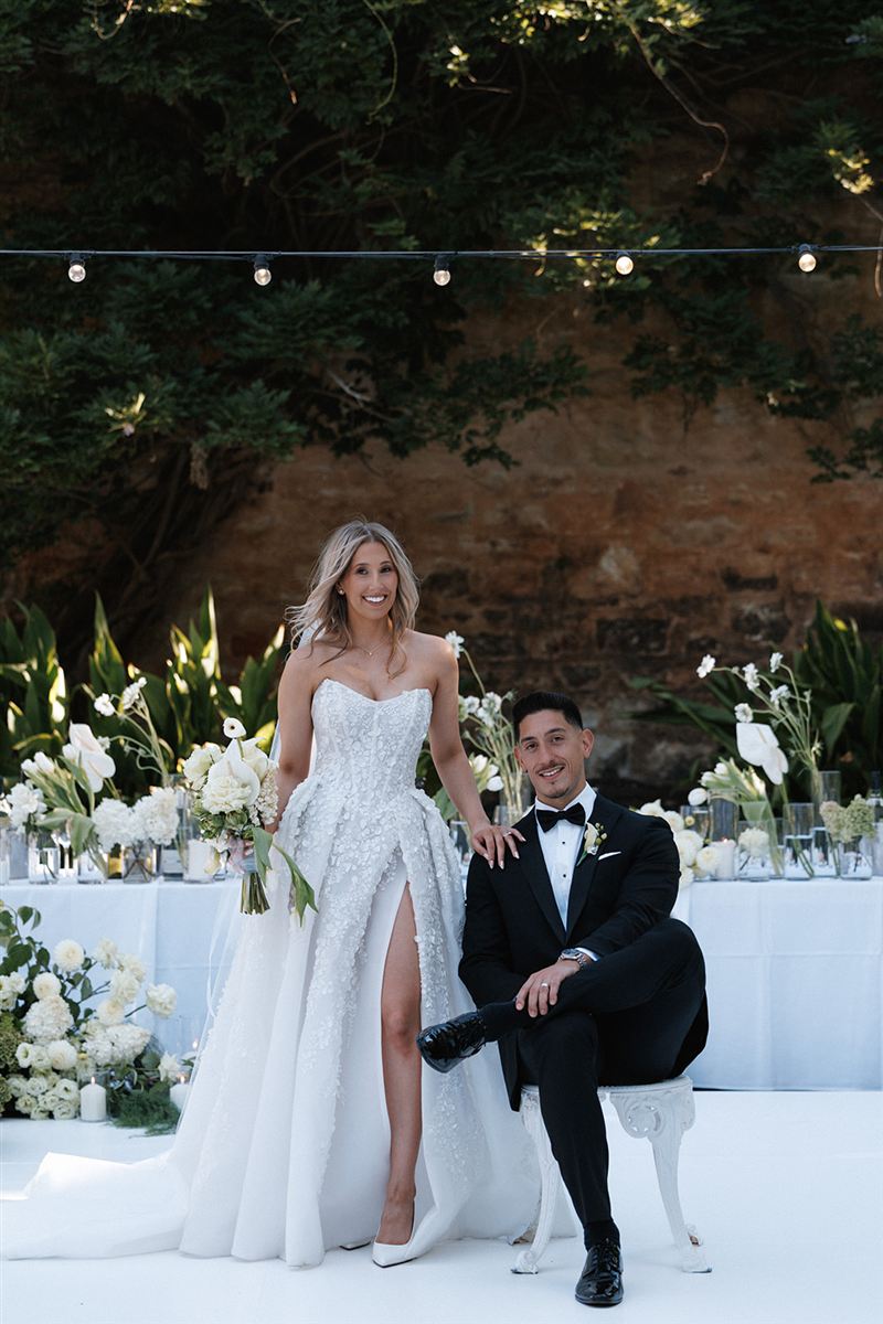Mia standing beside Luke, seated in front of their white floral reception table